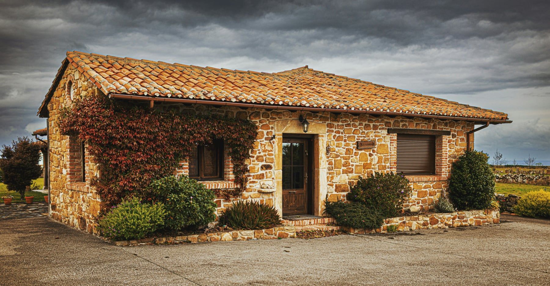 An ivy-clad Asturian stone cottage with a tiled roof, garden and cloudy sky